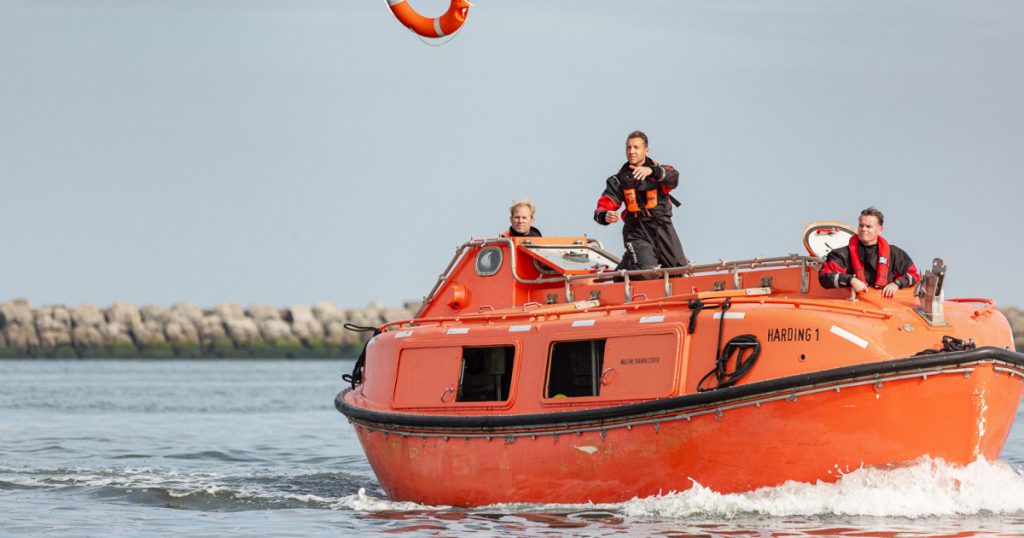 Un bote salvavidas de color naranja brillante con tres hombres a bordo. Uno de los hombres lanza un salvavidas al agua.